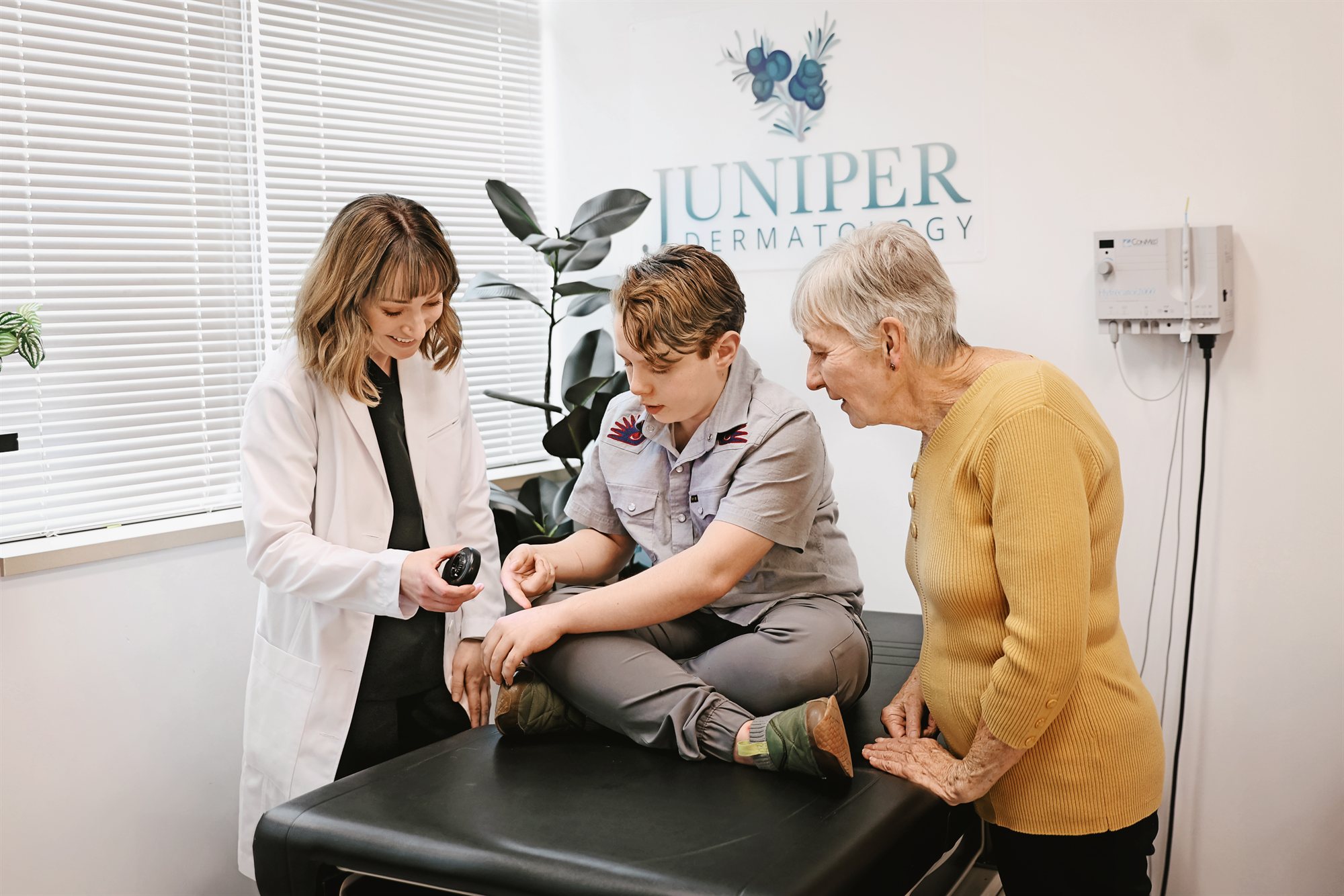 Elena Salpas, PA-C examining a young patient's arm with a dermatoscope while a family member watches at Juniper Dermatology